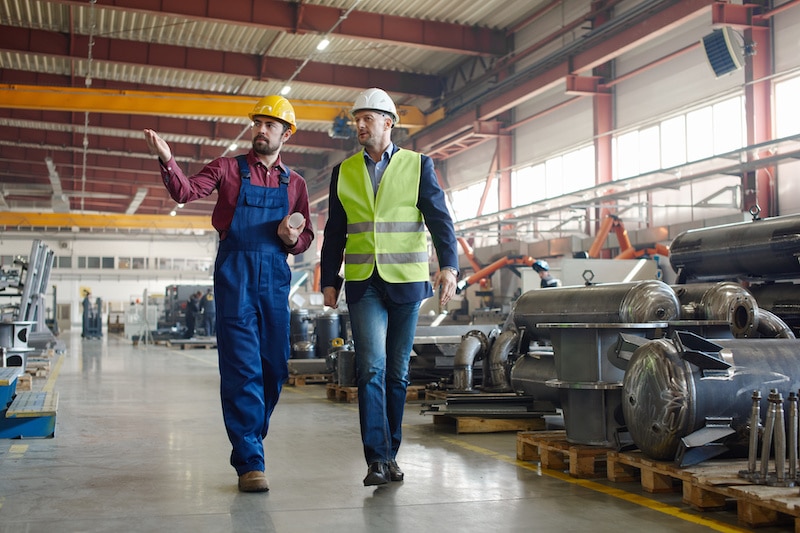 A view of two engineers in the uniform and helmets among the industrial equipment of the plant they are working in both talking to each other.